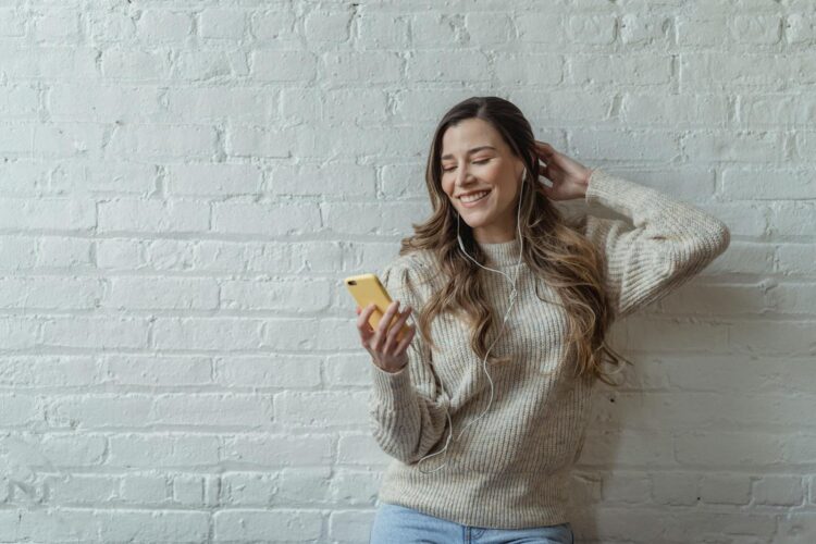 Smiling young woman using smartphone with earphones against a white brick wall.
